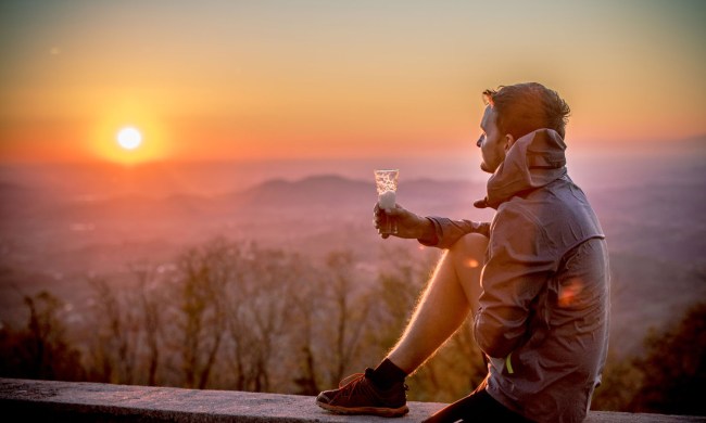 A man sitting on a wall and holding a glass of beer by the sunset.