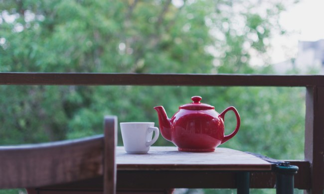 red teapot and white cup on wooden table outside with green trees in background